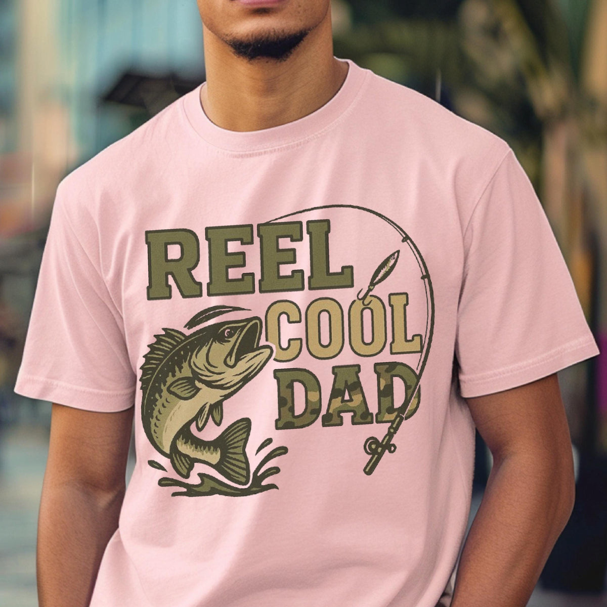 May include: A light pink short-sleeved t-shirt featuring a graphic print of a fish jumping out of water with a fishing rod and the text REEL COOL DAD in olive green and camouflage patterns. The design is centered on the front of the garment.