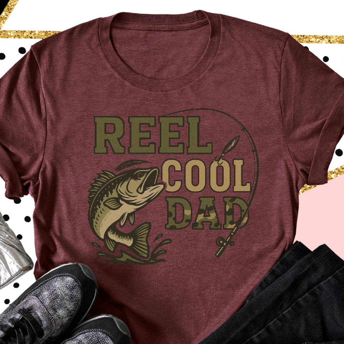 May include: A maroon short-sleeved t-shirt featuring a graphic print of a fish jumping out of water with a fishing rod and the text REEL COOL DAD in olive green and camouflage lettering. The design is centered on the front of the garment.