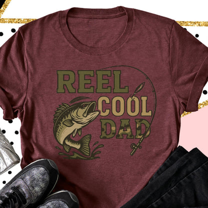 May include: A maroon short-sleeved t-shirt featuring a graphic print of a fish jumping out of water with a fishing rod and the text REEL COOL DAD in olive green and camouflage lettering. The design is centered on the front of the garment.