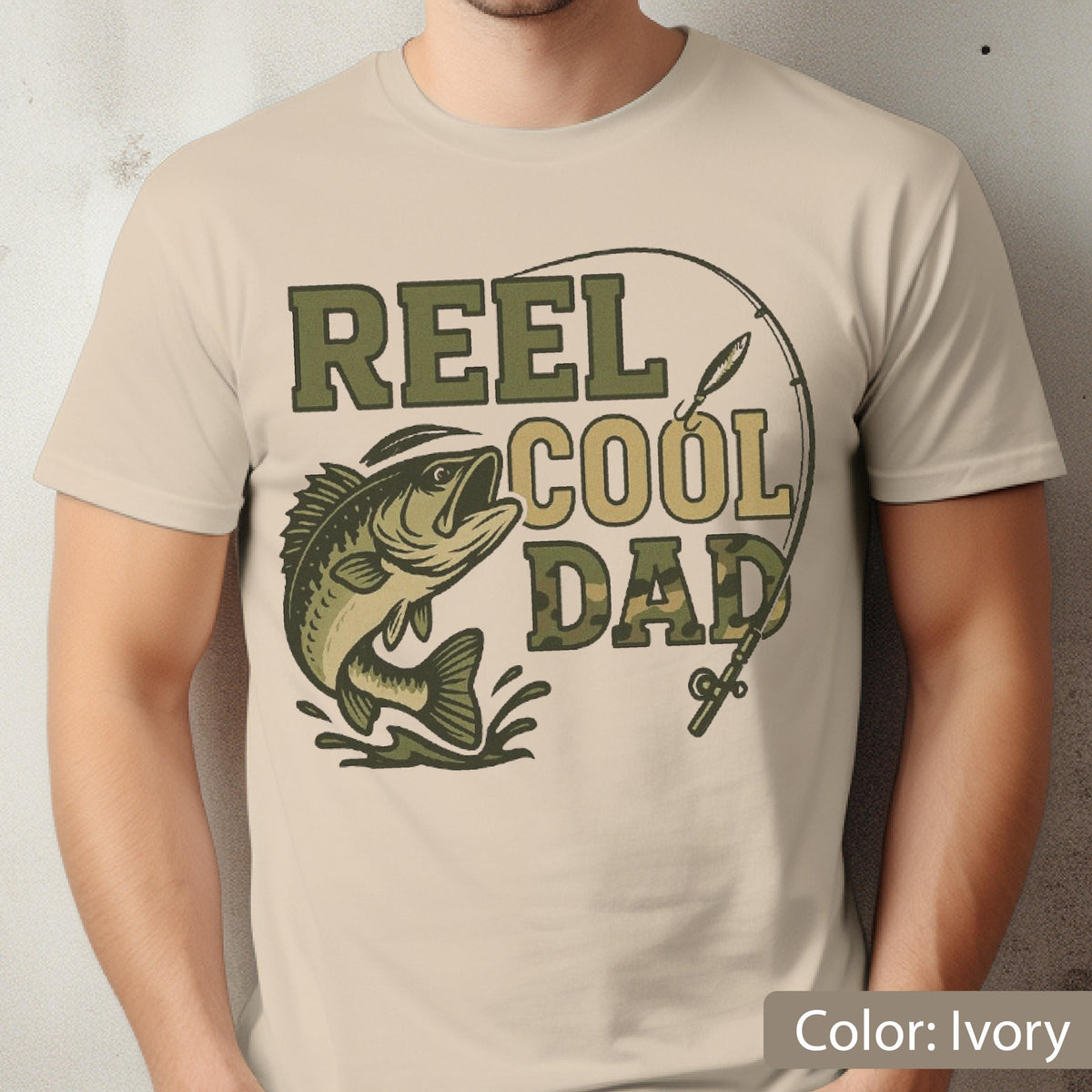May include: Ivory-colored t-shirt featuring a graphic with the text "REEL COOL DAD." The design includes a fish leaping from the water, a fishing rod, and text in shades of green and tan. The shirt is a short-sleeved crew neck.