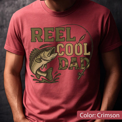 May include: Crimson t-shirt with the text "REEL COOL DAD" in green and tan. A graphic of a fish jumping out of the water is on the left. A fishing rod and hook are above the text. The shirt is a solid crimson color.
