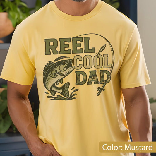 May include: Mustard yellow t-shirt featuring the text "REEL COOL DAD" in green and camouflage lettering, with a graphic of a fish and fishing rod. The shirt is short-sleeved.