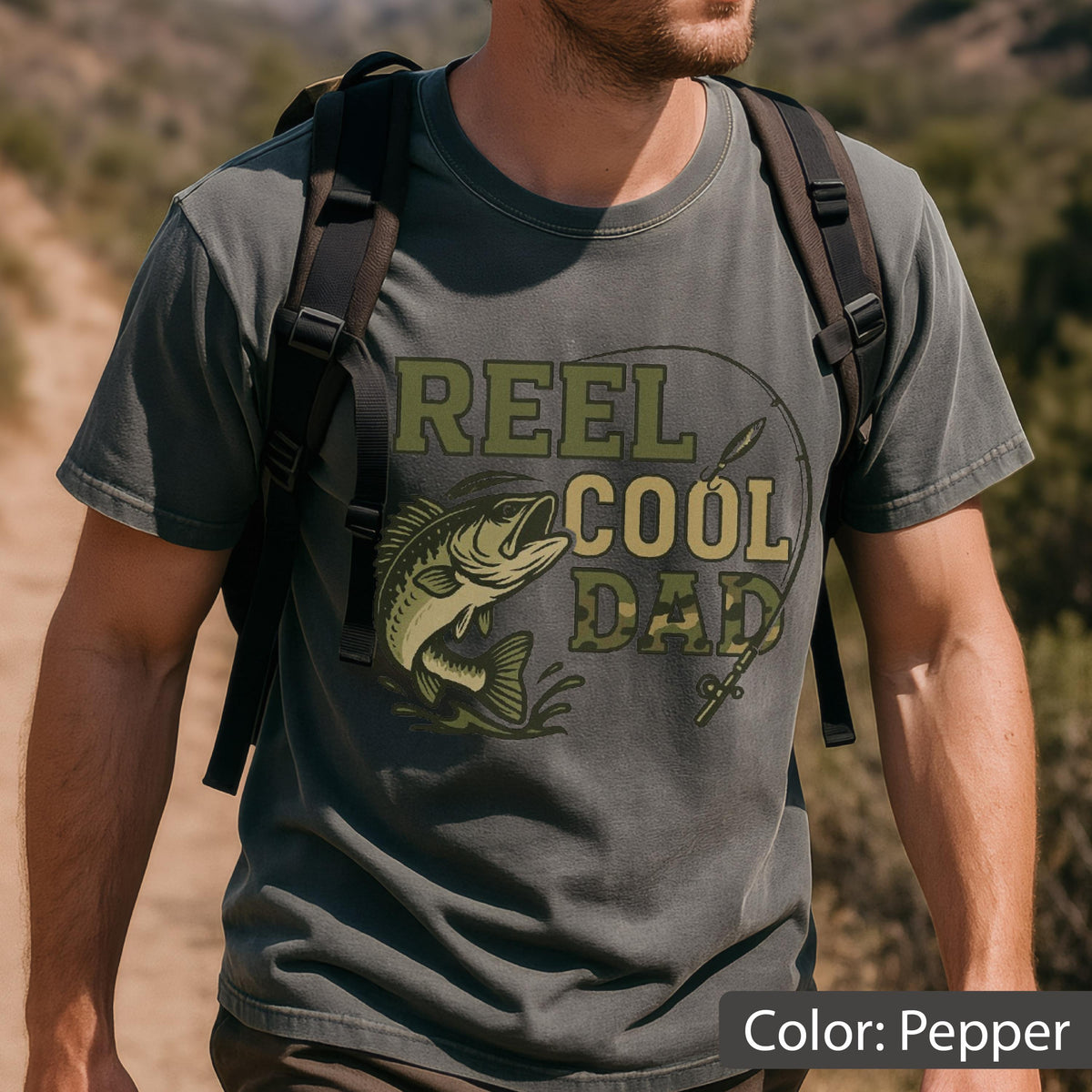 May include: Pepper-colored t-shirt with the text "REEL COOL DAD" in green and tan camouflage lettering. A graphic of a fish and fishing rod is also on the shirt. The shirt is short-sleeved.