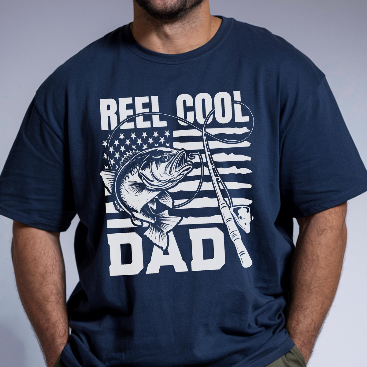 May include: Navy blue t-shirt with a graphic design featuring a fish, fishing rod, and the words "REEL COOL DAD" in white. The design incorporates an American flag motif. The shirt is a crew neck style.