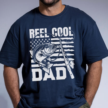 May include: Navy blue t-shirt with a graphic design featuring a fish, fishing rod, and the words "REEL COOL DAD" in white. The design incorporates an American flag motif. The shirt is a crew neck style.