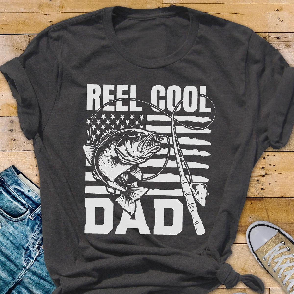 May include: Dark gray t-shirt with a white graphic featuring a fish, fishing rod, and the words "REEL COOL DAD." The design incorporates an American flag motif. The shirt is made of a soft material.