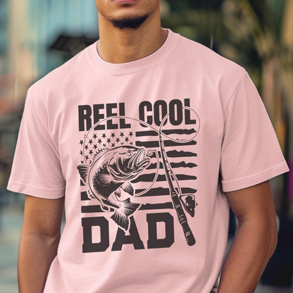 May include: A light pink t-shirt with a black graphic design. The design features the words "REEL COOL DAD" above and below an illustration of a fish and fishing rod, with an American flag background.