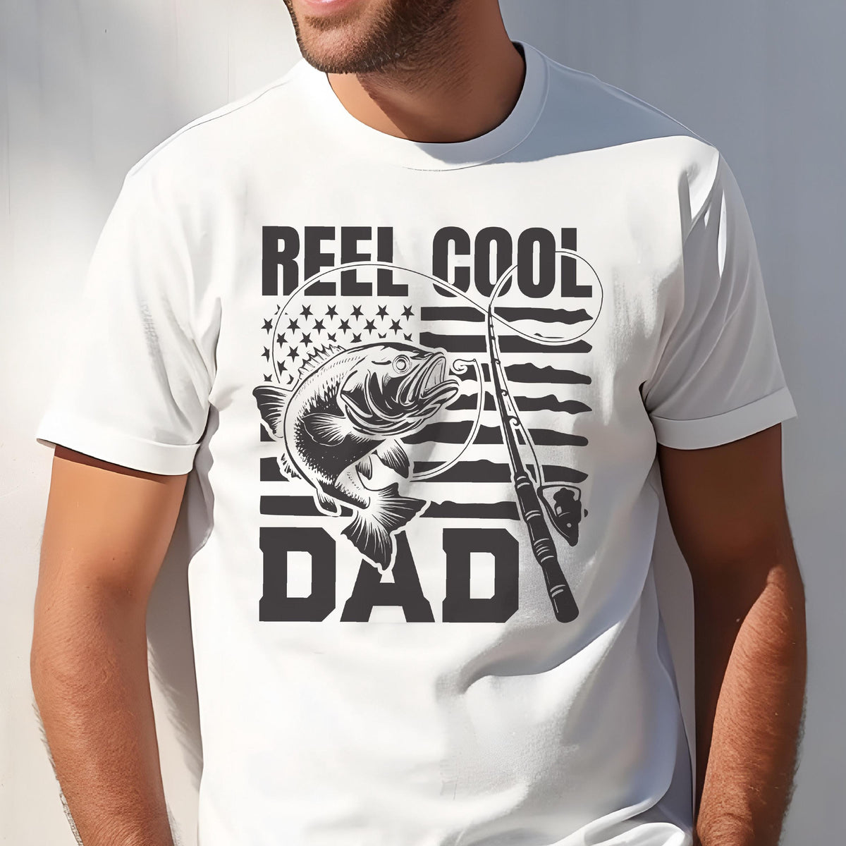 May include: White t-shirt with a black graphic featuring a fish, fishing rod, and the words "REEL COOL DAD." The design incorporates elements of the American flag. The shirt is a casual wear item.