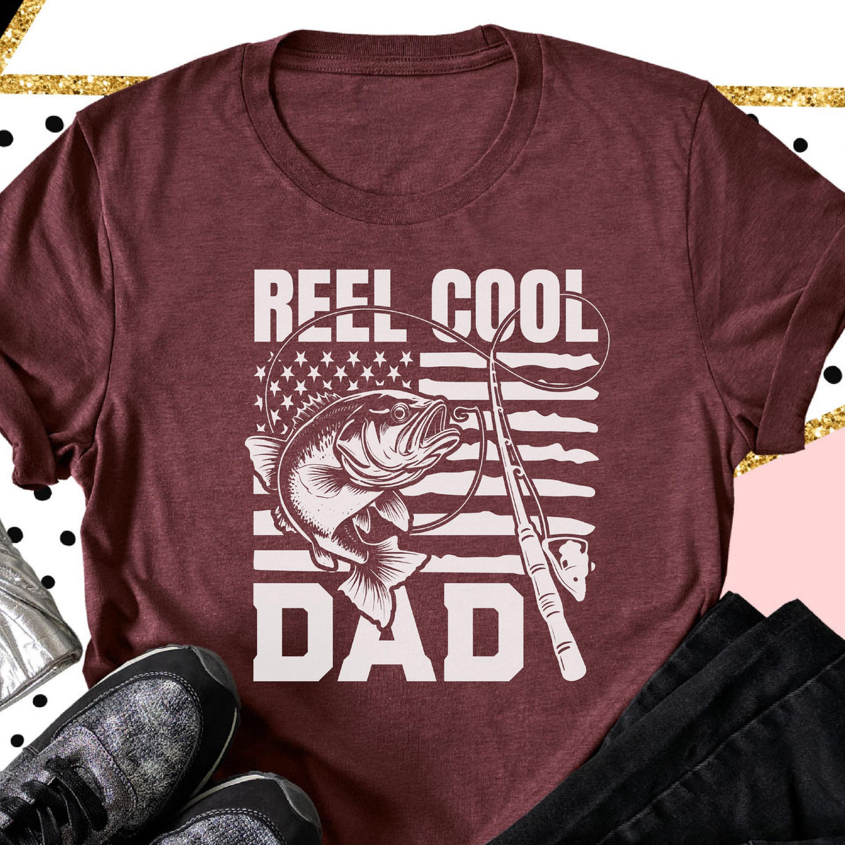 May include: Maroon t-shirt with a white graphic featuring a fish, fishing rod, and the words "REEL COOL DAD." The design incorporates the American flag with stars and stripes. The shirt has a crew neck and short sleeves.