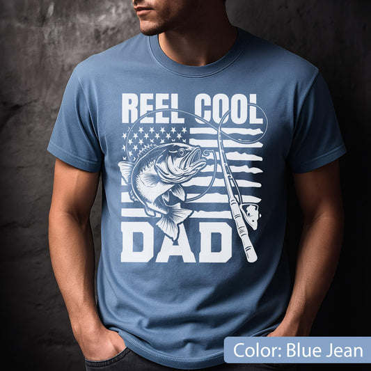 May include: Blue jean-colored t-shirt with a white graphic. The graphic features a fish, fishing rod, and the words "REEL COOL DAD" with an American flag design in the background. The shirt is short-sleeved.