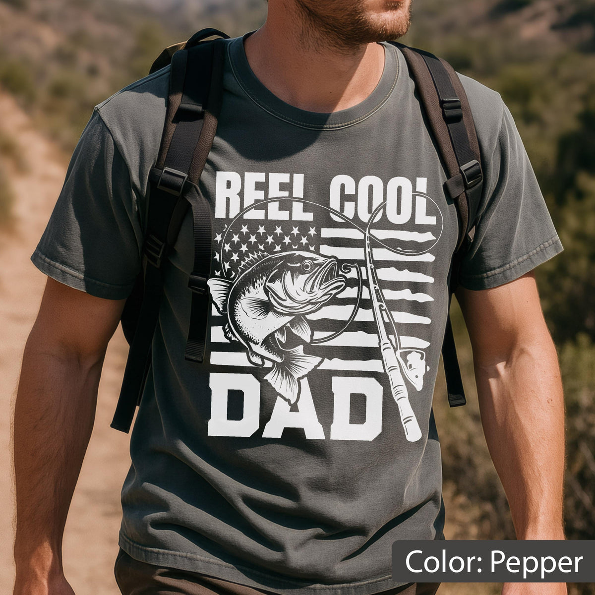 May include: Pepper-colored t-shirt with the text "REEL COOL DAD" in white. The design includes a fish, fishing rod, and an American flag motif. The shirt is a crew neck and has short sleeves.