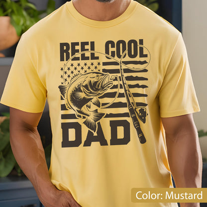 May include: Mustard yellow t-shirt with a black graphic. The graphic includes the text "REEL COOL DAD", a fish, a fishing rod, and a design resembling the American flag. The shirt is short-sleeved.