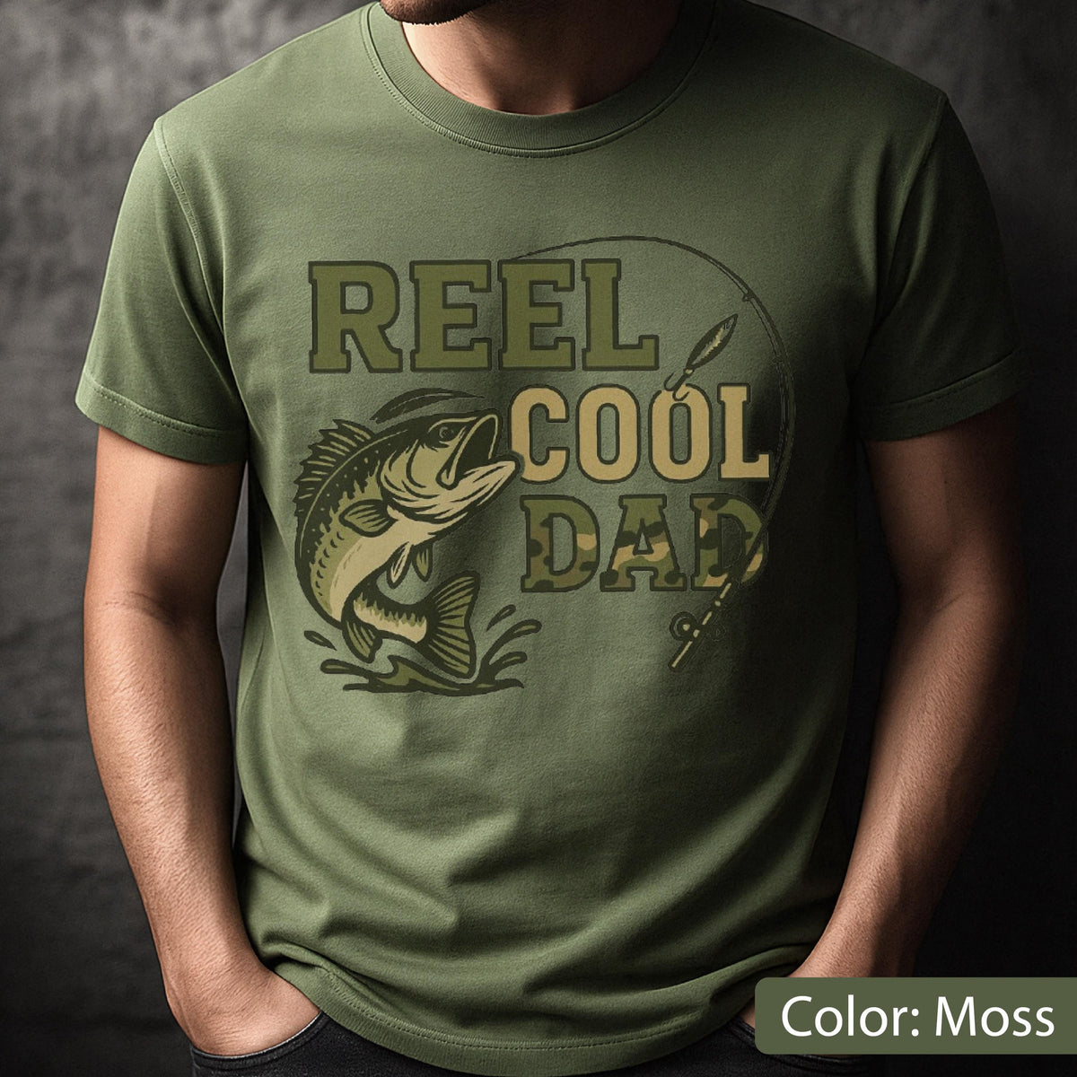 May include: Moss green t-shirt with a graphic design featuring a fish jumping out of water, a fishing rod, and the text "REEL COOL DAD" in shades of green and tan. The shirt is a short-sleeved crew neck.