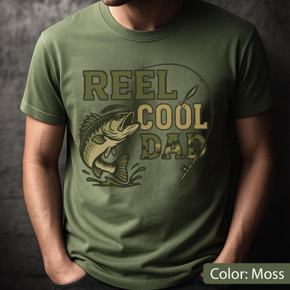 May include: Moss green t-shirt with a graphic design featuring a fish jumping out of water, a fishing rod, and the text "REEL COOL DAD" in shades of green and tan. The shirt is a short-sleeved crew neck.