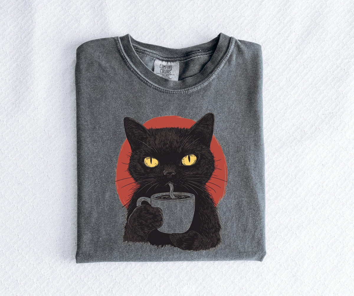 May include: A charcoal gray t-shirt featuring a graphic illustration of a black cat with bright yellow eyes holding a mug of coffee. Behind the cat is a solid red circle. The shirt has a crew neckline and a Comfort Colors brand label at the neck.