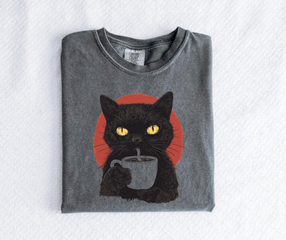 May include: A charcoal gray t-shirt featuring a graphic illustration of a black cat with bright yellow eyes holding a mug of coffee. Behind the cat is a solid red circle. The shirt has a crew neckline and a Comfort Colors brand label at the neck.