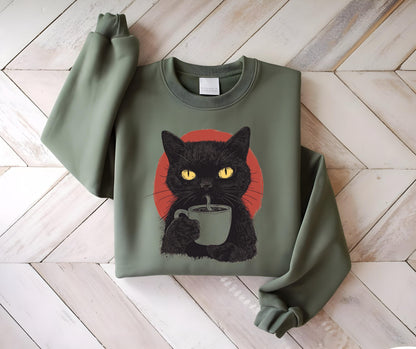 May include: An olive green crewneck sweatshirt featuring a graphic print of a black cat with bright yellow eyes holding a light gray mug. Behind the cat is a solid red circle. The sweatshirt has long sleeves and a ribbed collar and cuffs.