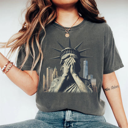May include: Dark gray t-shirt featuring a distressed graphic of the Statue of Liberty covering her face, with a New York City skyline in the background. The shirt has a vintage, worn look.