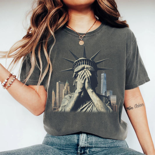 May include: Dark gray t-shirt featuring a distressed graphic of the Statue of Liberty covering her face, with a New York City skyline in the background. The shirt has a vintage, worn look.
