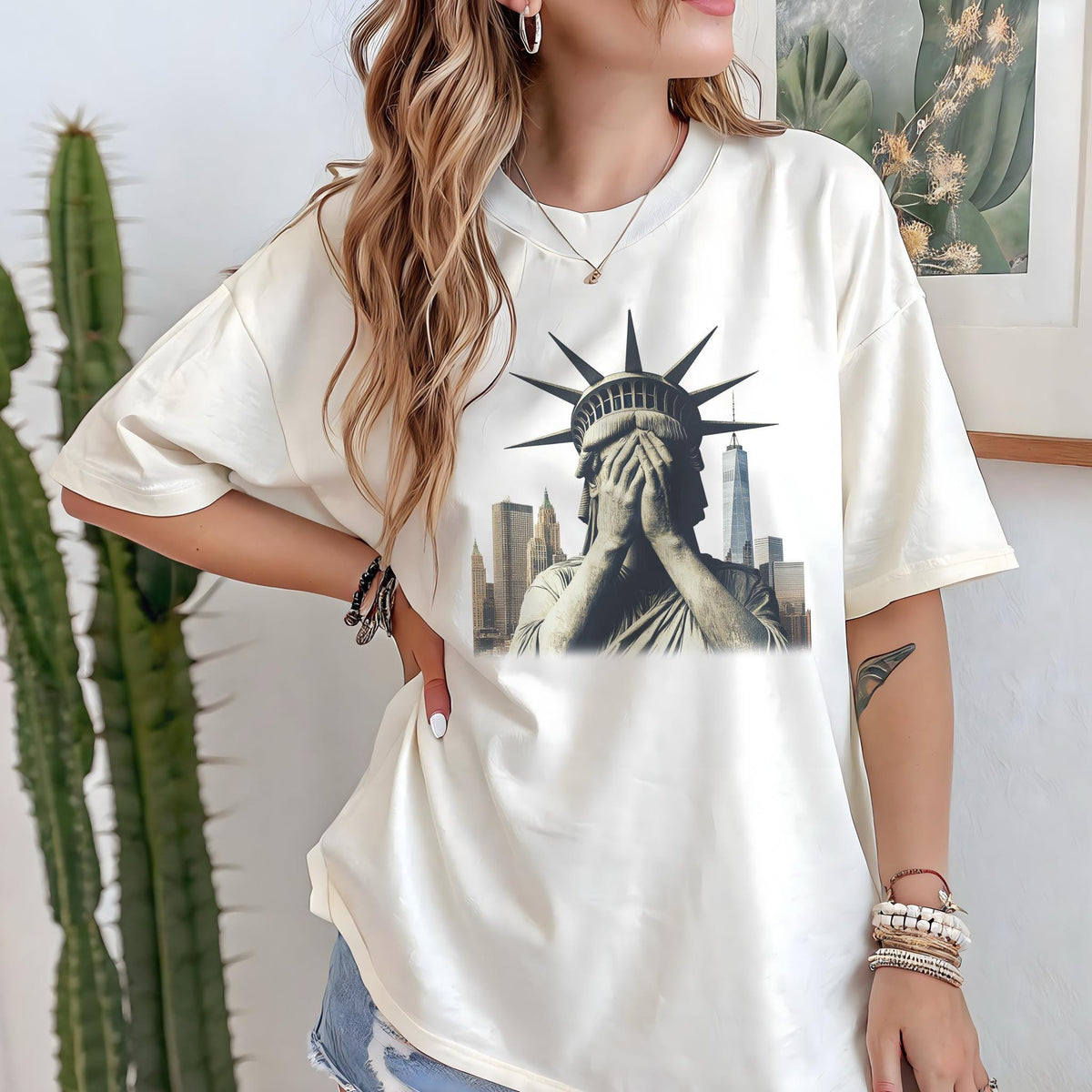 May include: Cream-colored t-shirt featuring a graphic of the Statue of Liberty with hands covering her face, set against a New York City skyline. The shirt has short sleeves and a relaxed fit.