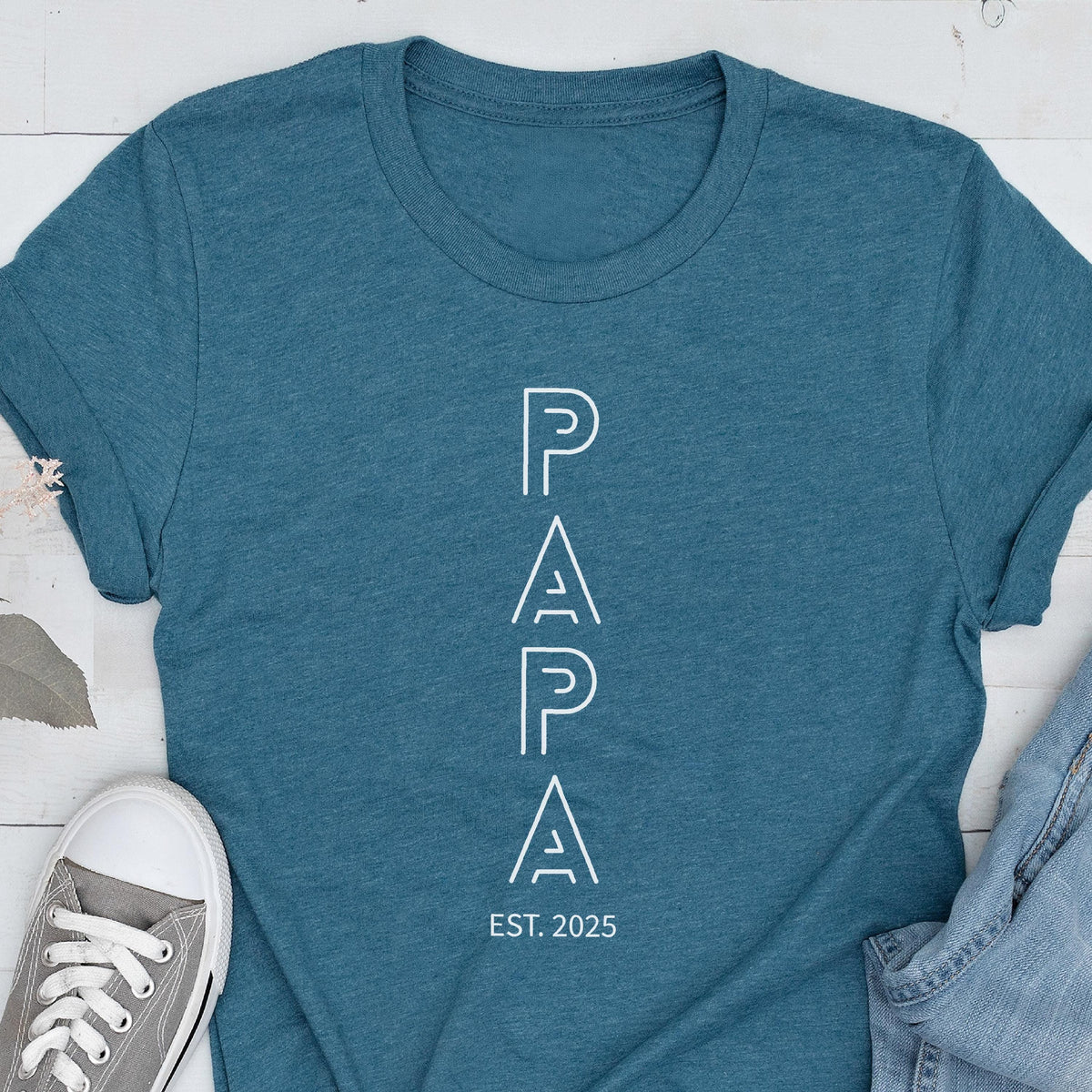 May include: Teal t-shirt with the word "PAPA" in white, vertical, outlined letters. Below the word "PAPA" is the text "EST. 2025". The shirt is made of a soft material and is suitable for casual wear.