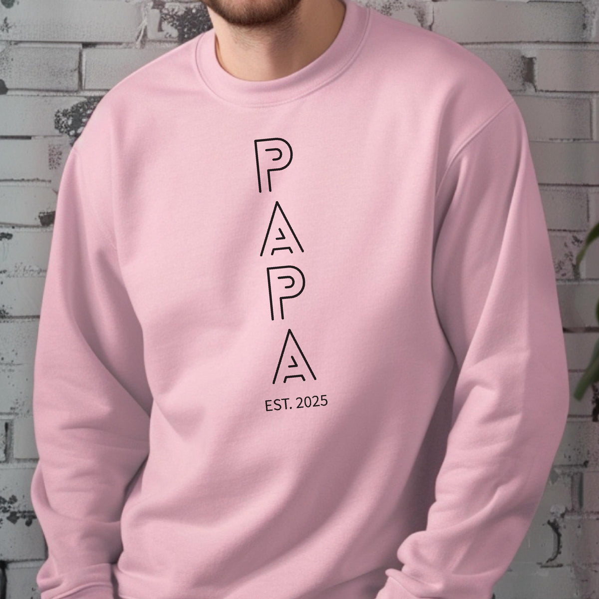 May include: A light pink sweatshirt with the word "PAPA" printed vertically in black, with "EST. 2025" below. The sweatshirt is a solid color and has a crew neck.
