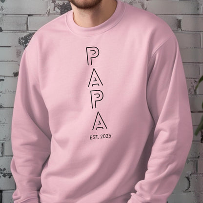 May include: A light pink sweatshirt with the word "PAPA" printed vertically in black, with "EST. 2025" below. The sweatshirt is a solid color and has a crew neck.