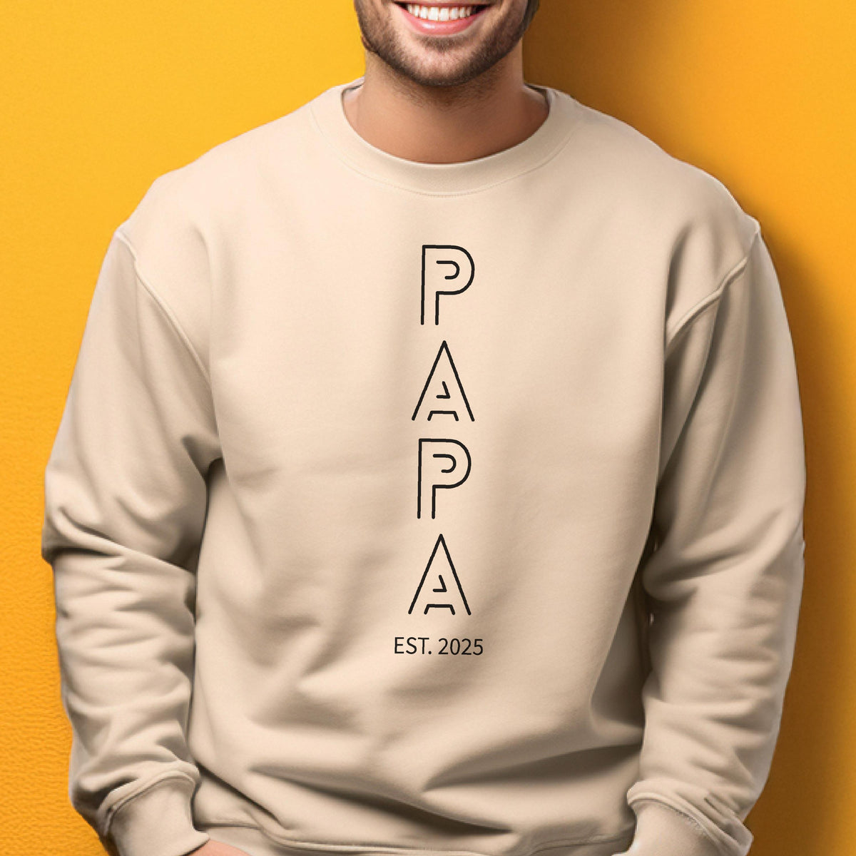 May include: A beige sweatshirt with the word "PAPA" in black, stylized, vertical letters. Below the word "PAPA" is the text "EST. 2025". The sweatshirt is a neutral color and has a crew neck.