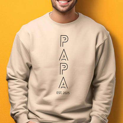 May include: A beige sweatshirt with the word "PAPA" in black, stylized, vertical letters. Below the word "PAPA" is the text "EST. 2025". The sweatshirt is a neutral color and has a crew neck.