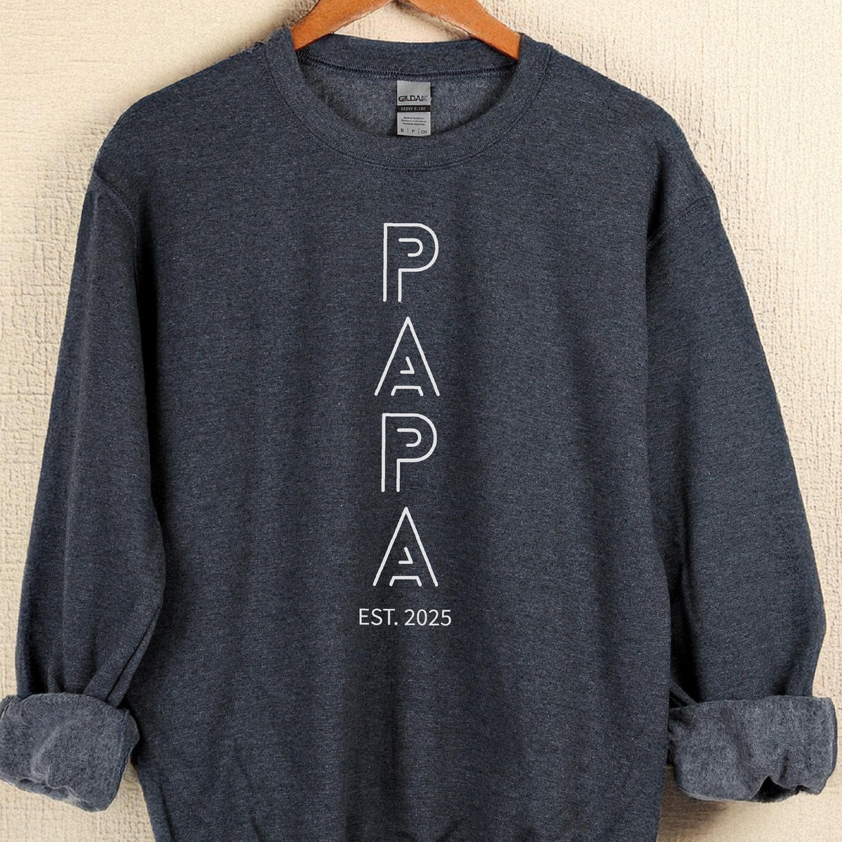 May include: A dark gray sweatshirt with the word "PAPA" in white, outlined letters. Below the word "PAPA" is the text "EST. 2025". The sweatshirt has a crew neck and long sleeves with rolled-up cuffs.