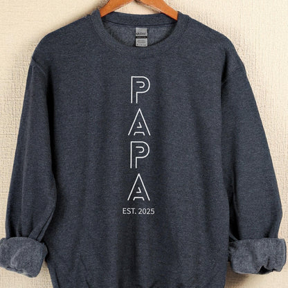 May include: A dark gray sweatshirt with the word "PAPA" in white, outlined letters. Below the word "PAPA" is the text "EST. 2025". The sweatshirt has a crew neck and long sleeves with rolled-up cuffs.
