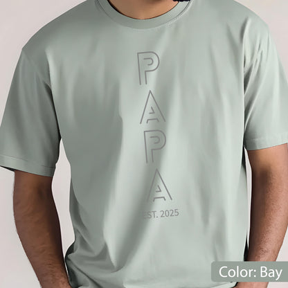 May include: A light green t-shirt with the word "PAPA" vertically printed in a lighter shade of green. Below the word "PAPA" is the text "EST. 2025". The shirt is a solid color and has short sleeves.
