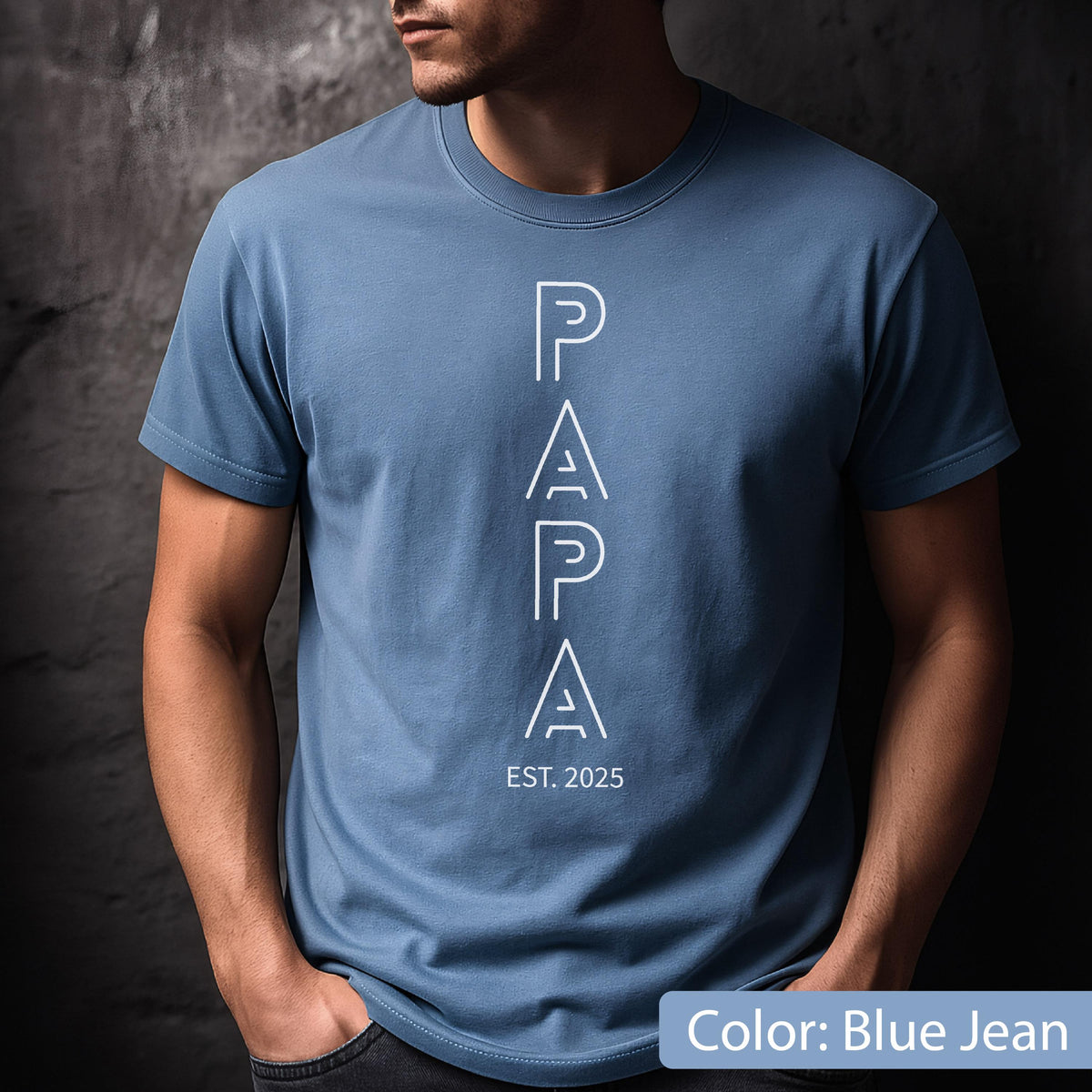 May include: Blue jean-colored t-shirt with the word "PAPA" in white, vertically aligned. Below the word "PAPA" is the text "EST. 2025". The shirt has short sleeves and a crew neck.