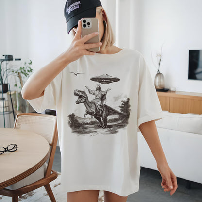 May include: Cream-colored t-shirt featuring a black and white graphic of a figure riding a dinosaur with a UFO above. The design includes a landscape and a bird in flight. The shirt has a crew neck and short sleeves.