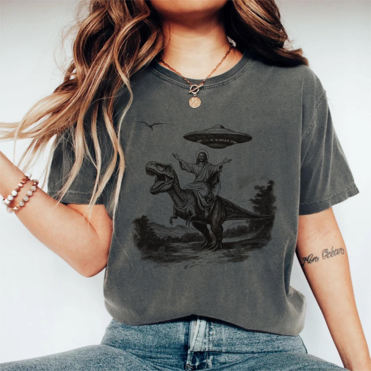 May include: Dark gray t-shirt featuring a graphic of Jesus riding a Tyrannosaurus Rex, with a UFO hovering above. The design is in black and white, with a landscape in the background. The shirt is a casual style.