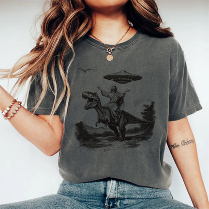 May include: Dark gray t-shirt featuring a graphic of Jesus riding a Tyrannosaurus Rex, with a UFO hovering above. The design is in black and white, with a landscape in the background. The shirt is a casual style.