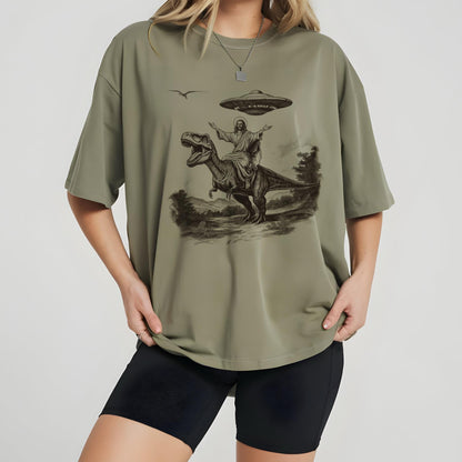 May include: Olive green t-shirt featuring a graphic of Jesus riding a Tyrannosaurus Rex with a UFO above. The design is in black and white, set against a landscape background. The shirt has short sleeves.