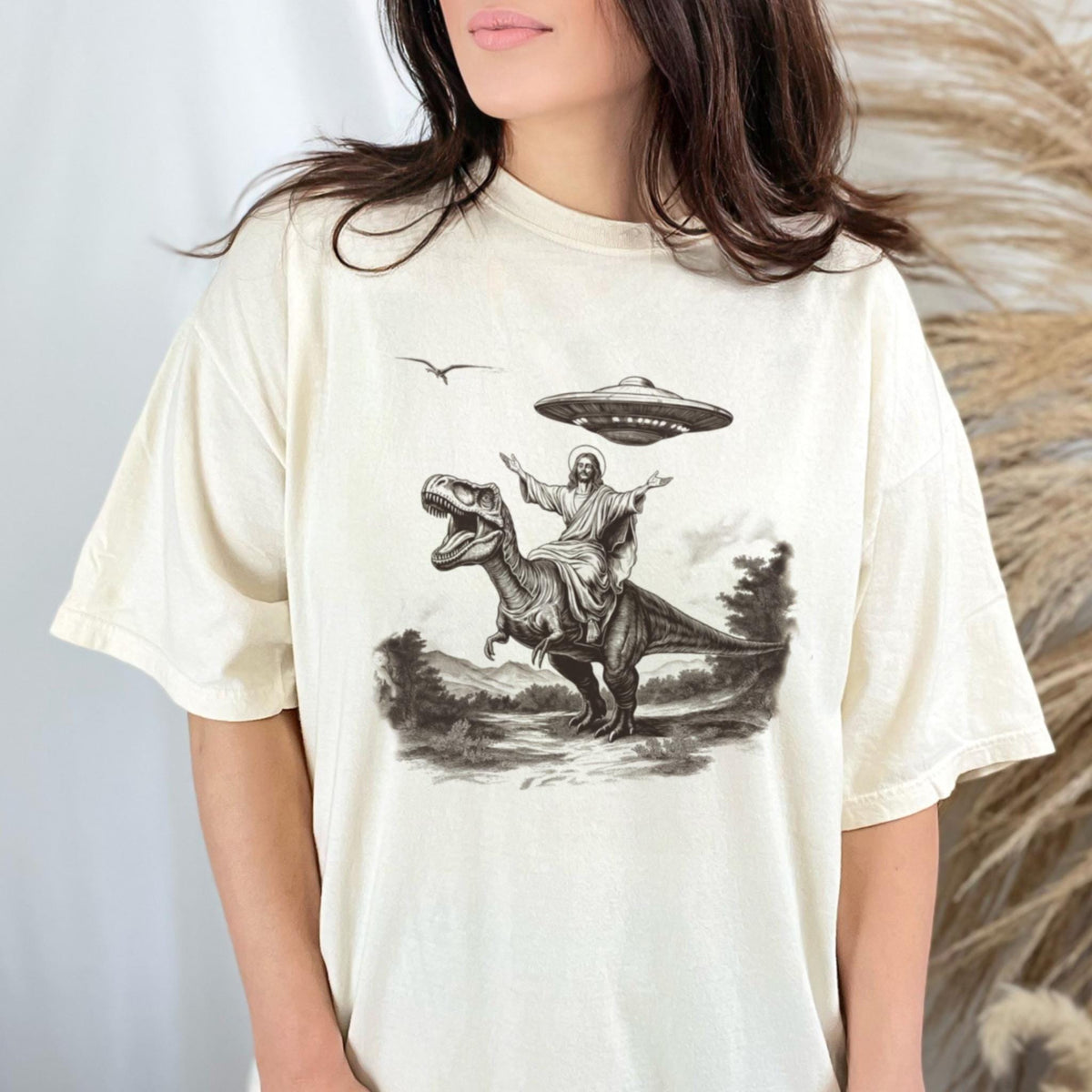 May include: Cream-colored t-shirt featuring a black and white graphic of Jesus riding a Tyrannosaurus Rex, with a UFO hovering above. The design has a vintage, distressed look. A bird flies in the background. The shirt is a casual, unisex style.