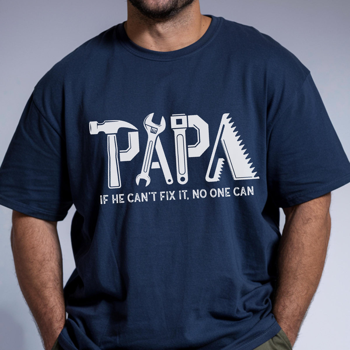 May include: Navy blue t-shirt with the word "PAPA" in white, stylized with tools. Below the word is the phrase "IF HE CAN'T FIX IT, NO ONE CAN". The shirt is a crew neck.