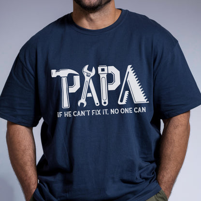 May include: Navy blue t-shirt with the word "PAPA" in white, stylized with tools. Below the word is the phrase "IF HE CAN'T FIX IT, NO ONE CAN". The shirt is a crew neck.