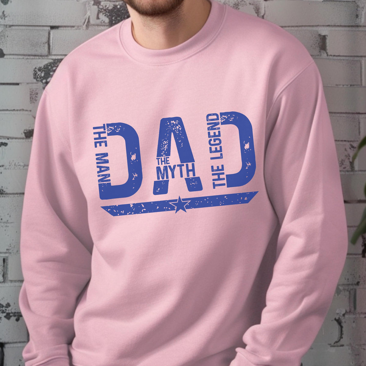 May include: A light pink sweatshirt with the words "DAD" in large, distressed blue letters. The text "THE MAN THE MYTH THE LEGEND" is arranged around the word "DAD", with a star graphic below. A casual, comfortable garment.