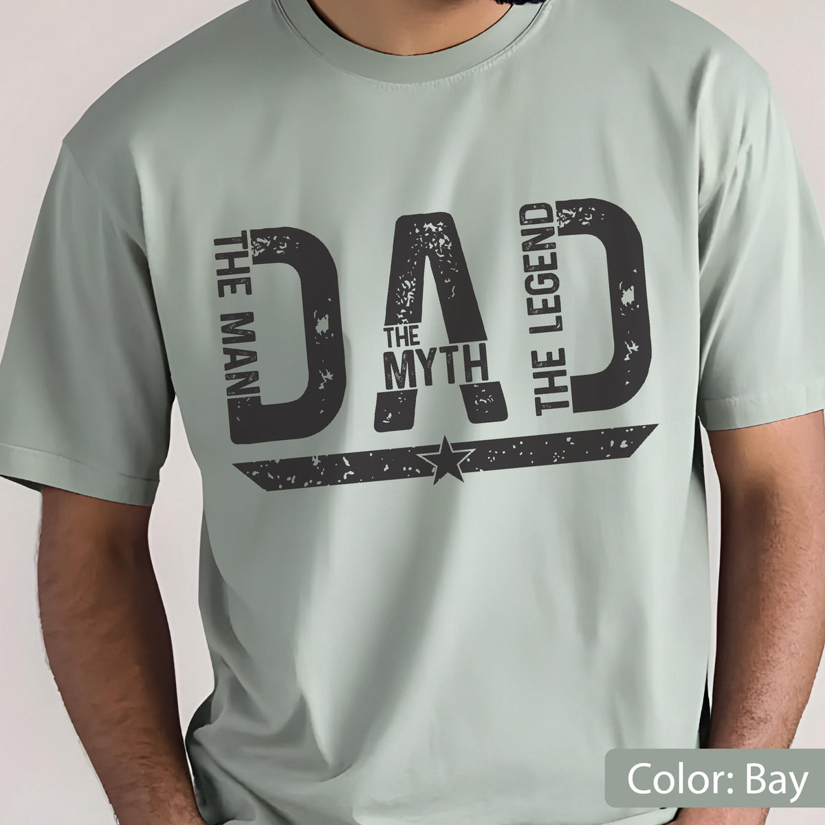 May include: A light green t-shirt with the word "DAD" in large, distressed black letters. The text "THE MAN THE MYTH THE LEGEND" is arranged around the word "DAD". A star graphic is below the word "MYTH".