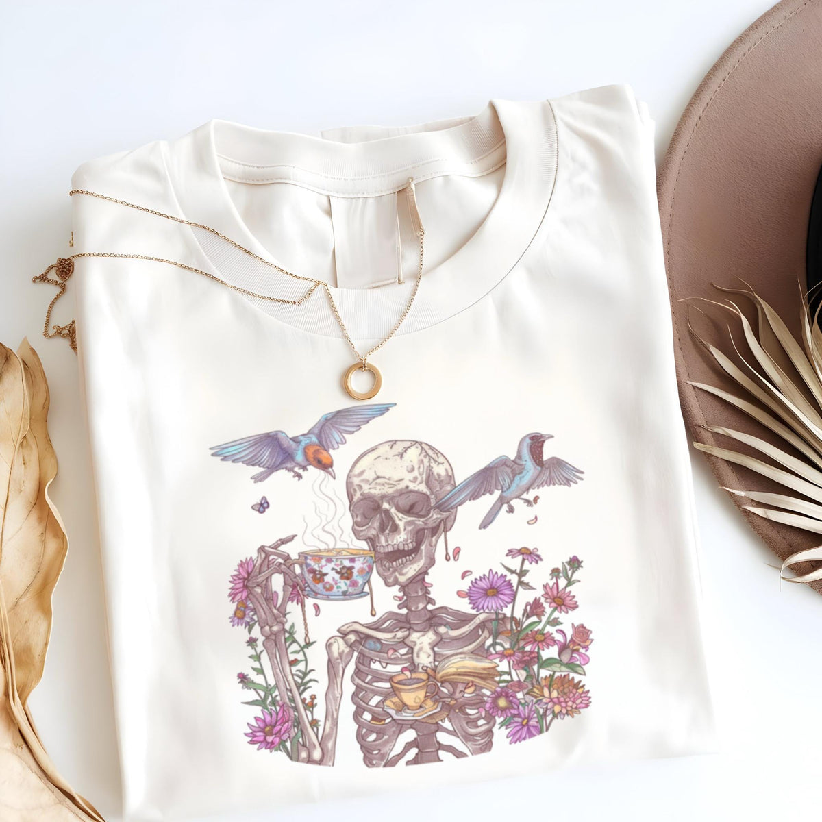 May include: Cream-colored t-shirt featuring a skeleton enjoying tea, surrounded by flowers and birds. The skeleton is holding a teacup and saucer. A gold necklace is draped over the shirt. The design is whimsical and artistic.