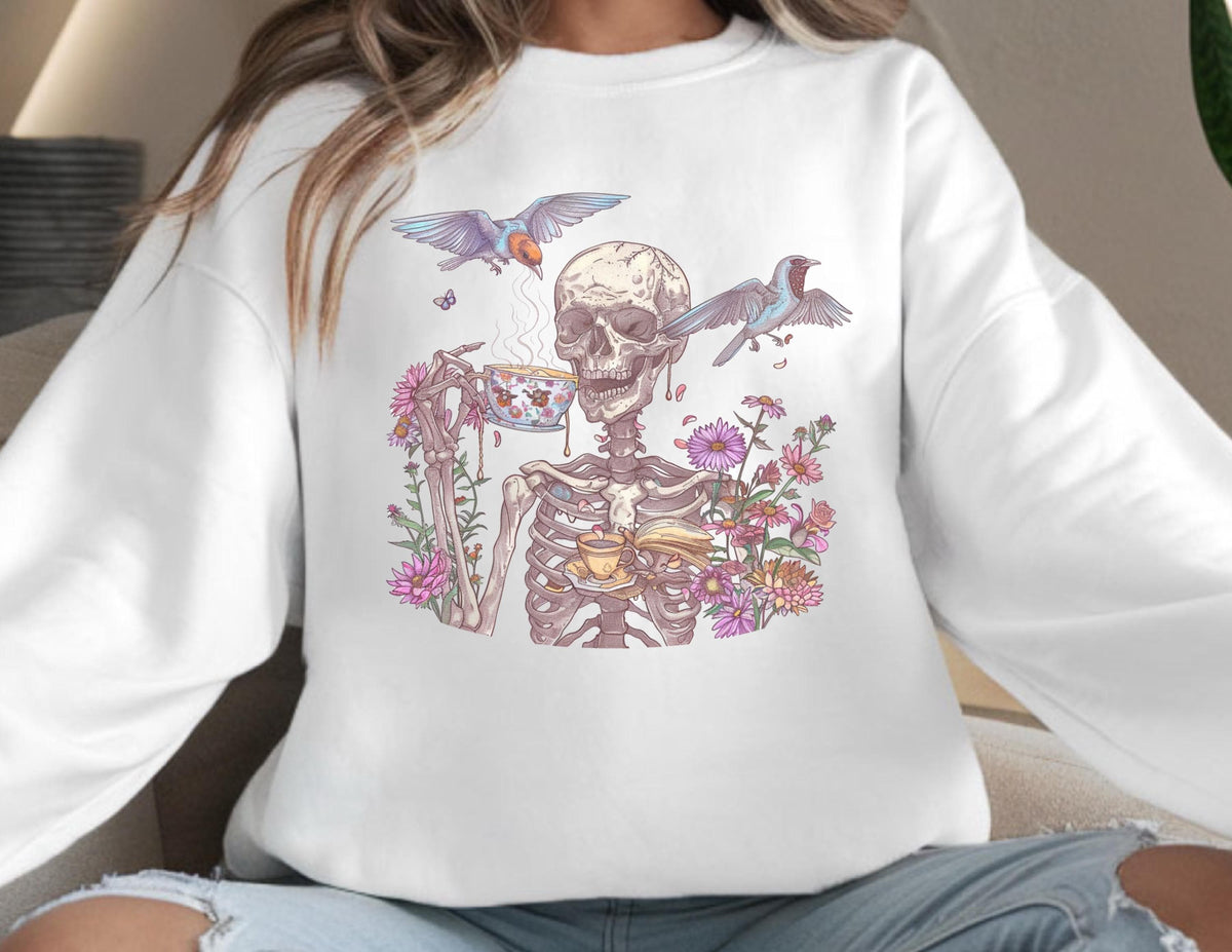 May include: White sweatshirt featuring a whimsical illustration of a skeleton enjoying tea, surrounded by flowers and birds. The design includes a teacup, a book, and steam rising from the cup, creating a unique and artistic aesthetic.