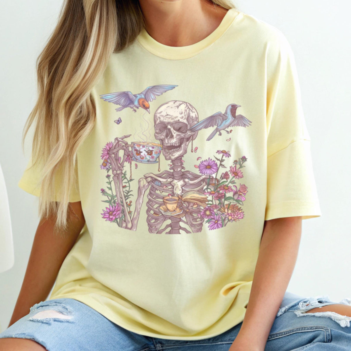 May include: A light yellow t-shirt featuring a skeleton enjoying tea, surrounded by flowers and birds. The skeleton is holding a teacup and saucer, with steam rising from the cup. The graphic is detailed and colorful.