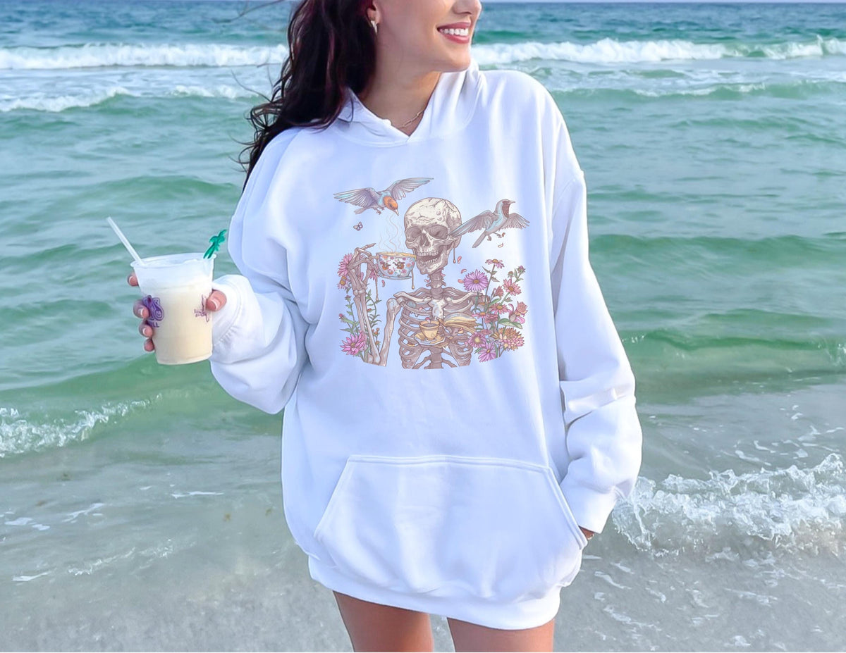 May include: White hoodie with a front pocket and a graphic of a skeleton with flowers and birds. The skeleton design is in shades of beige, pink, and green. The hoodie is worn on a beach with the ocean in the background.