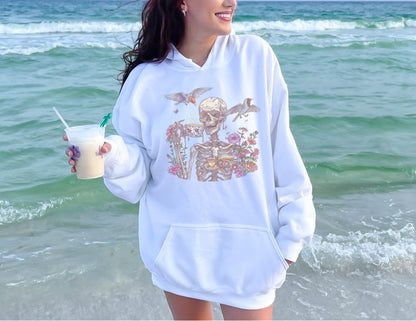 May include: White hoodie with a front pocket and a graphic of a skeleton with flowers and birds. The skeleton design is in shades of beige, pink, and green. The hoodie is worn on a beach with the ocean in the background.