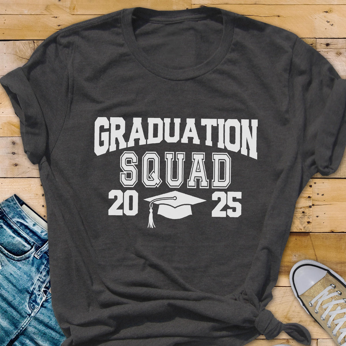 May include: Dark gray t-shirt with the words "GRADUATION SQUAD 2025" in white block letters. A white graduation cap is centered below the word "SQUAD". The shirt is a crew neck style.