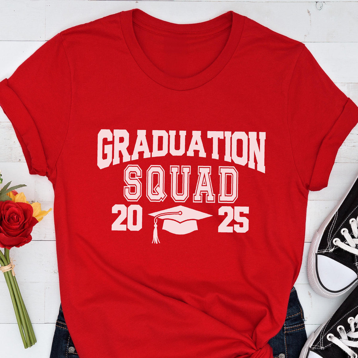 May include: A red t-shirt with the words "GRADUATION SQUAD 2025" in white block letters. A white graduation cap graphic is centered below the word "SQUAD". The shirt is folded at the sleeves.