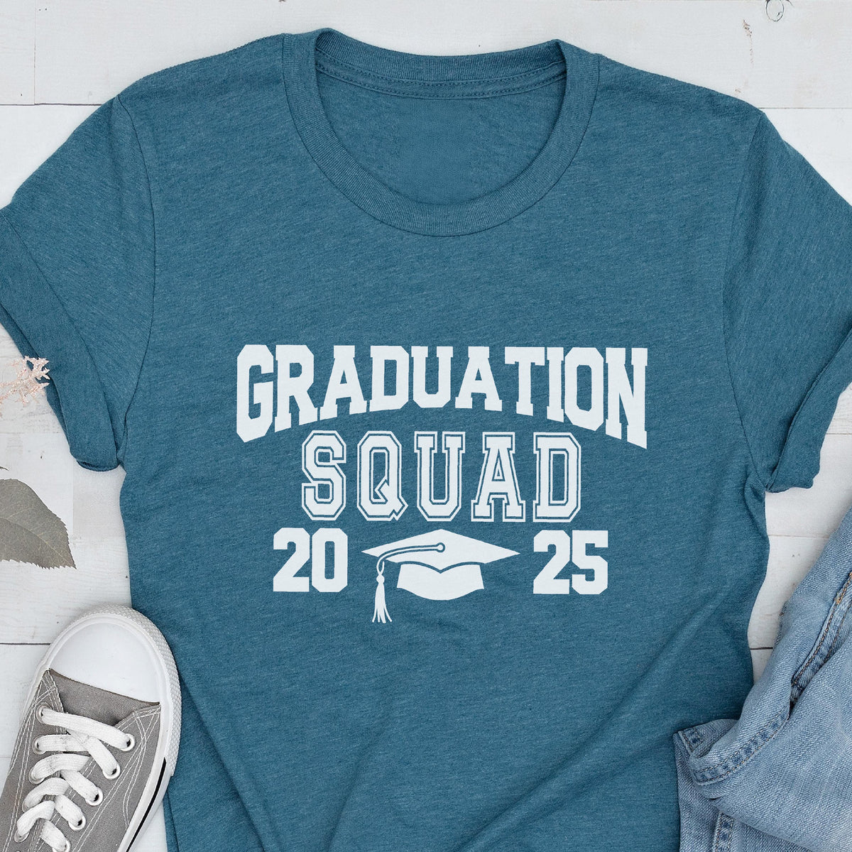 May include: Teal t-shirt with the words "GRADUATION SQUAD 2025" in white block letters. A graduation cap graphic is between the numbers. The shirt is paired with gray sneakers and blue jeans.